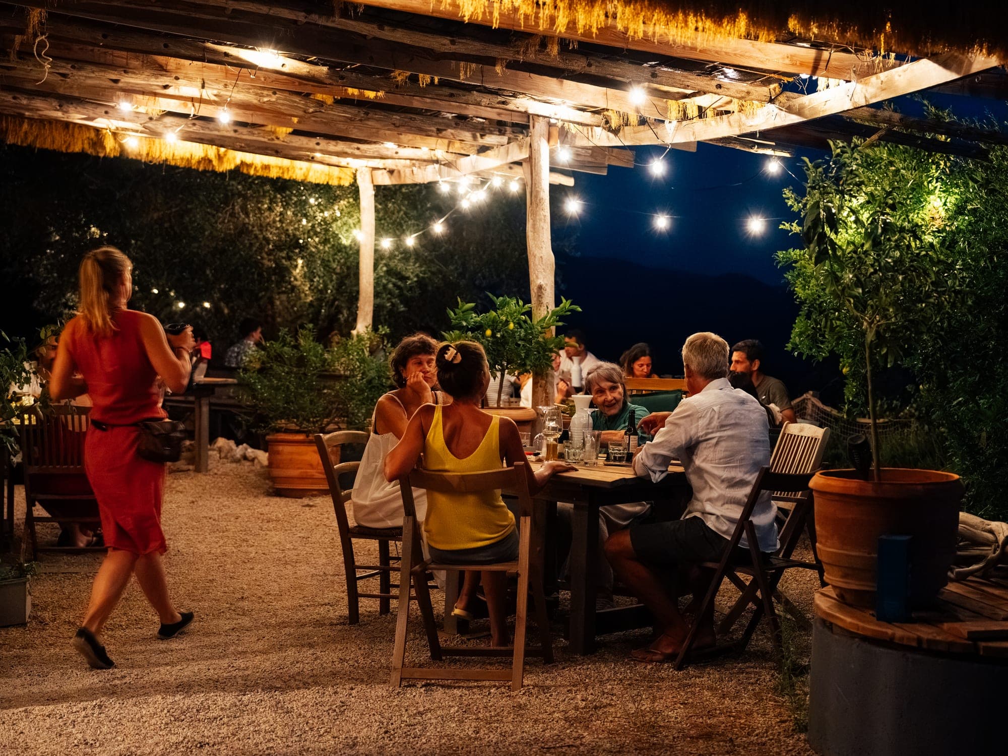 Gäste beim Abendessen an Holztischen unter der Pergola, darüber Lichterketten, dazwischen Zitronenbäumchen in Terrakottatöpfen