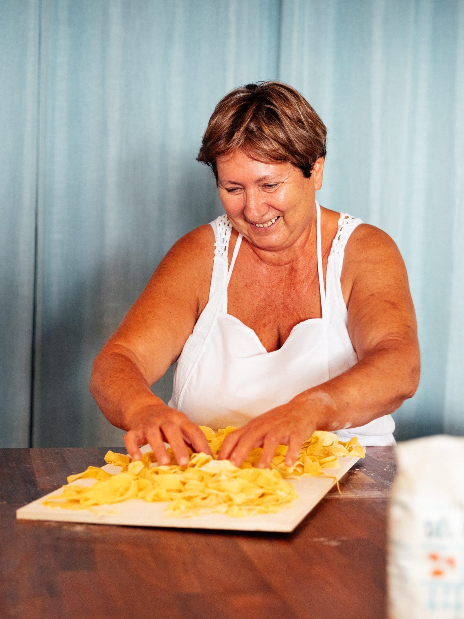 Eine Frau in weißer Schürze lockert lachend frisch geschnittene Pappardelle auf einem Holzbrett