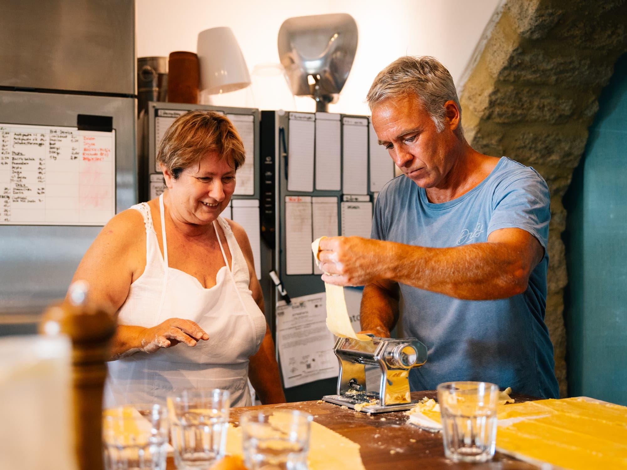Eine Frau in weißer Schürze und ein Mann an der Nudelmaschine, er zieht eine frische Teigbahn durch