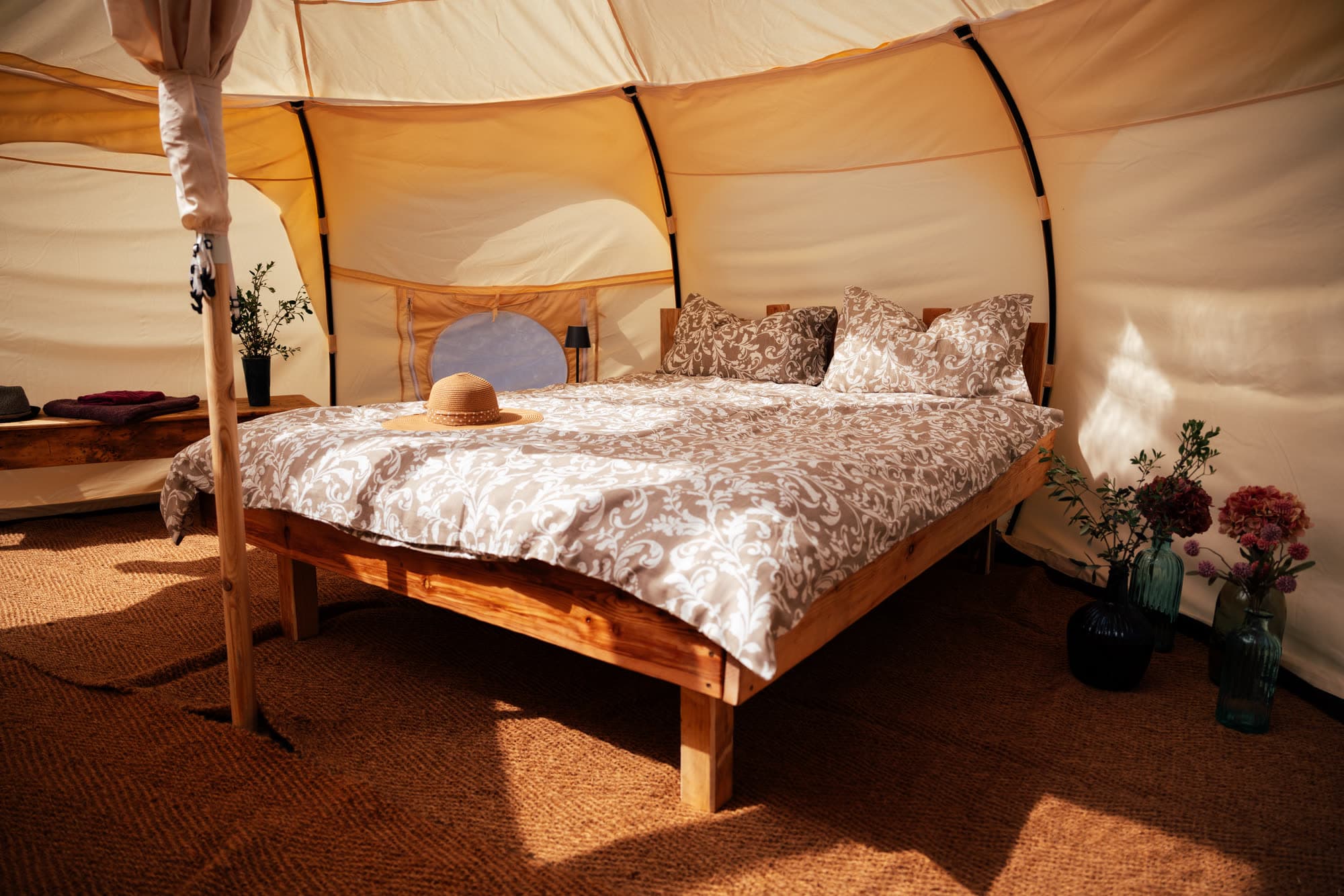 Wooden double bed with patterned bedding and a straw hat, beside it flowers in glass bottles and a round tent window