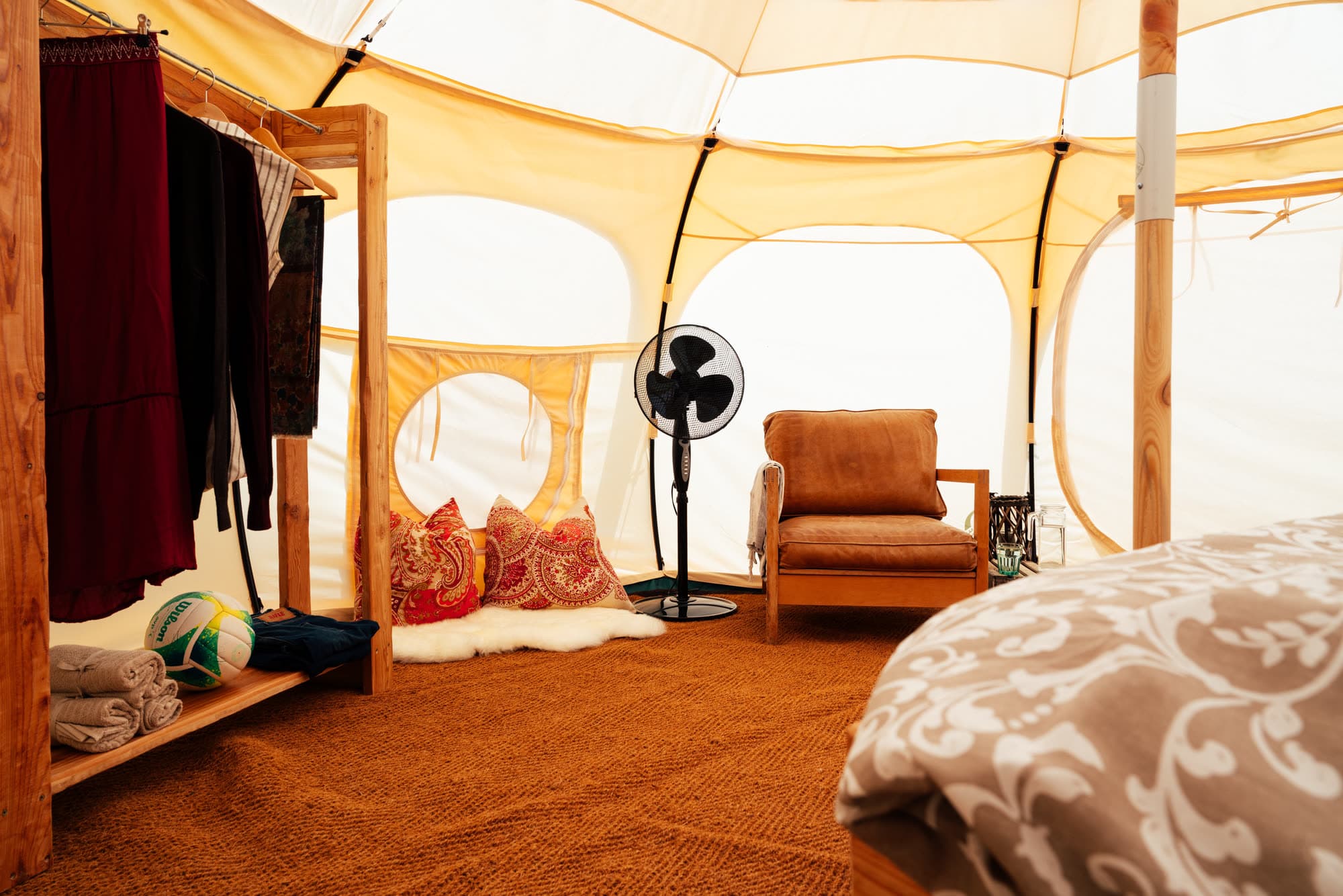 Across the bell tent: a clothes rail with towels, paisley cushions on a sheepskin and a leather armchair by the wooden pole