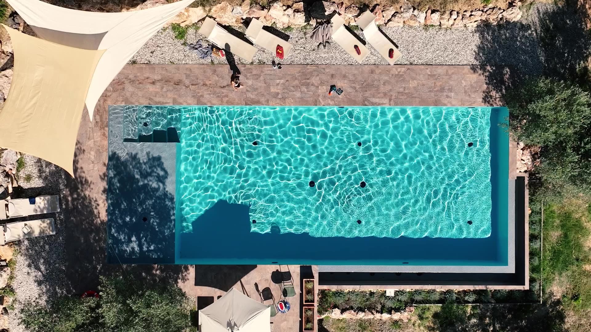 Pool auf einer Terrasse, dahinter eine Trockenmauer mit Liegen, Sonnensegel und Olivenbäume