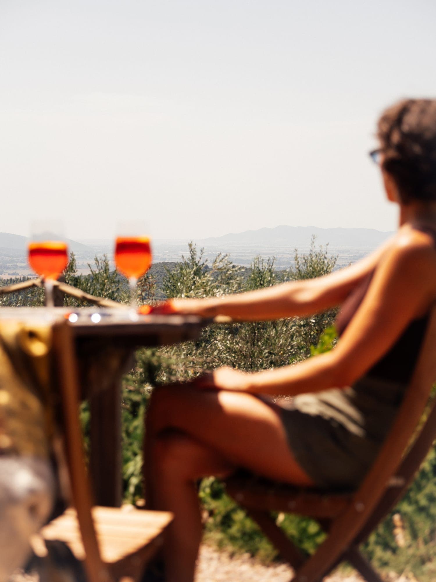 En gæst i en liggestol med to Aperol-glas på bordet og udsigt ud over olivenskråningerne til Maremmas bakker