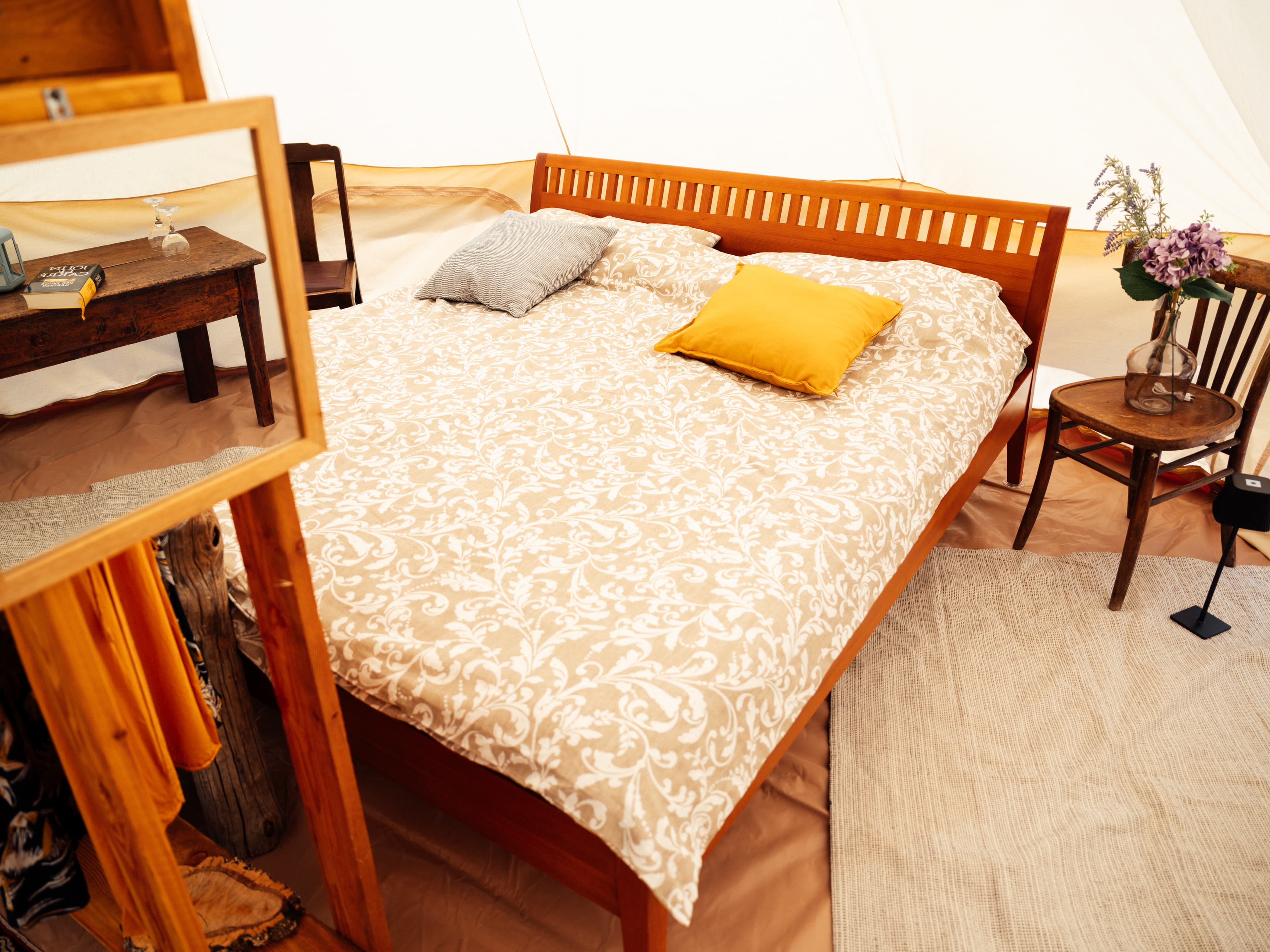 Double bed with a beige patterned throw and a yellow cushion, beside it a chair with a vase of flowers