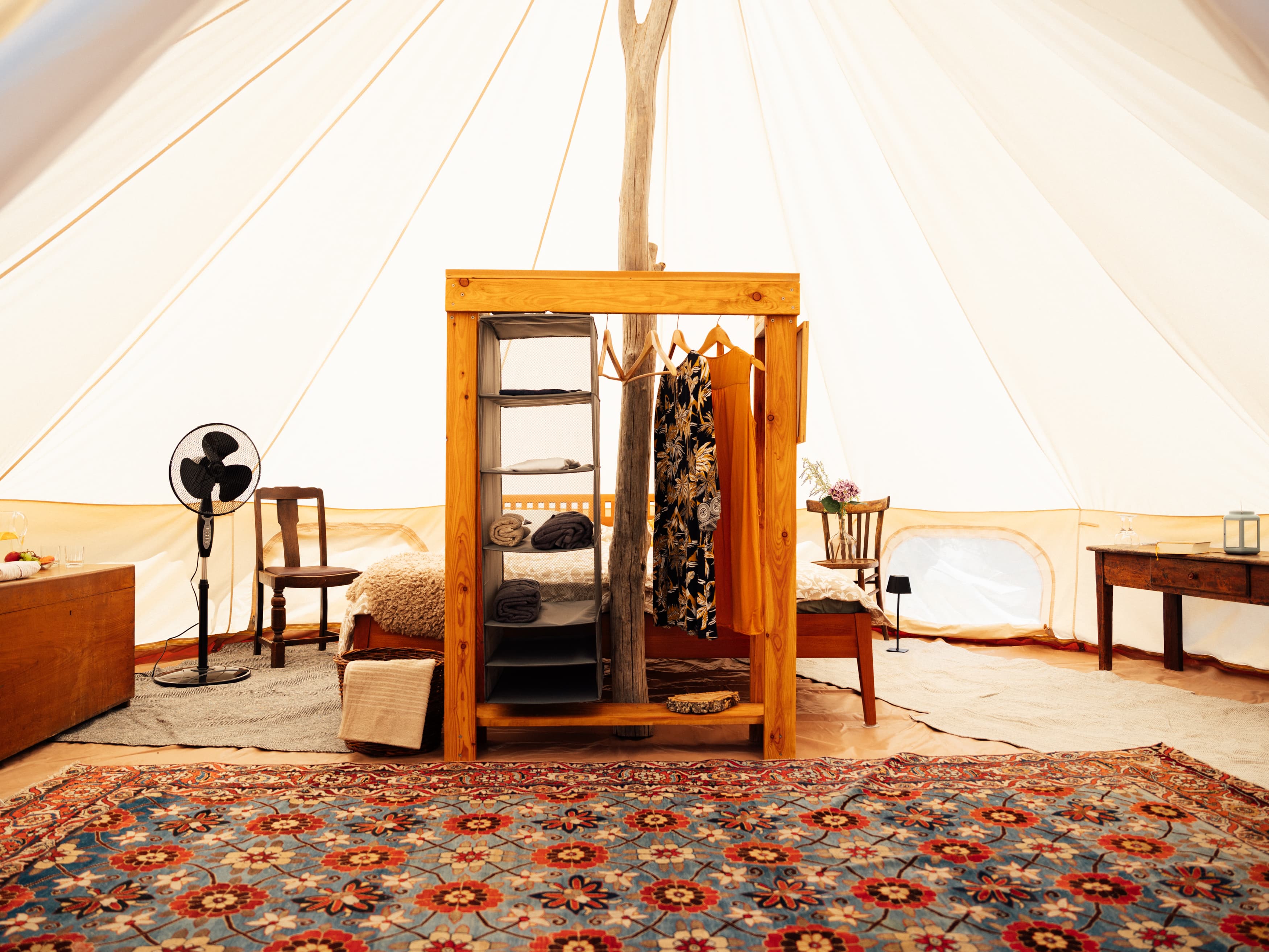 Tent interior with an open wooden wardrobe, a fan, the bed and a floral rug
