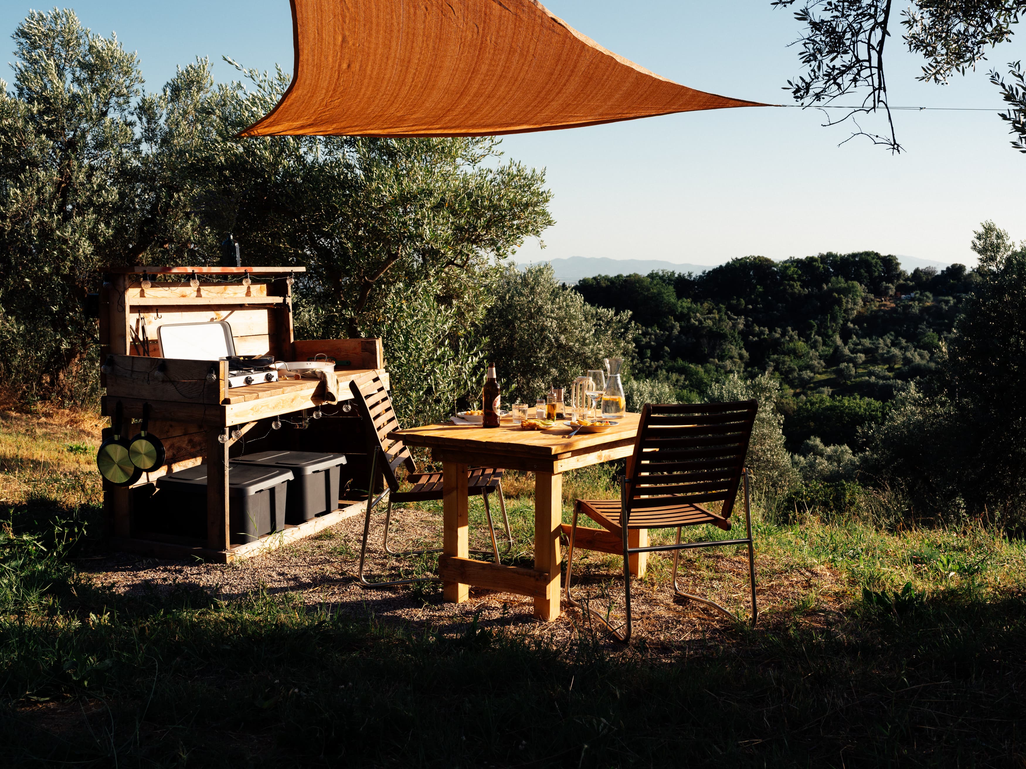 Wooden outdoor kitchen with a gas stove under a shade sail, beside it a dining table looking out over the valley