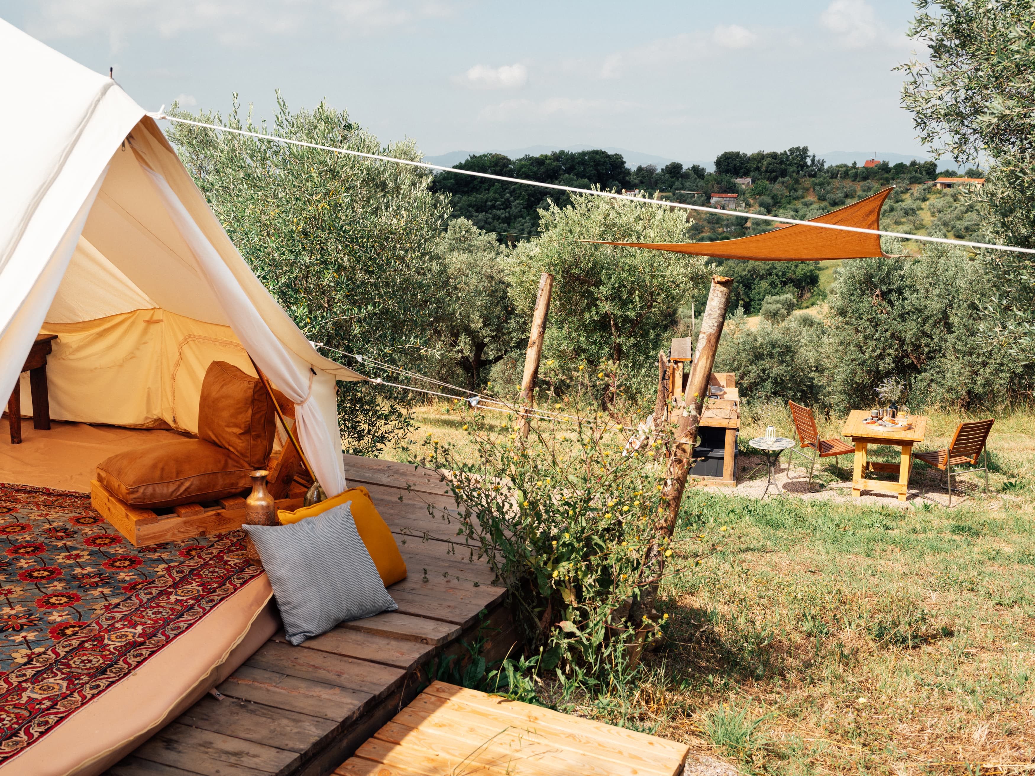 Large Sahara tent with a wooden deck and cushions, beside it an outdoor kitchen and dining table under a shade sail