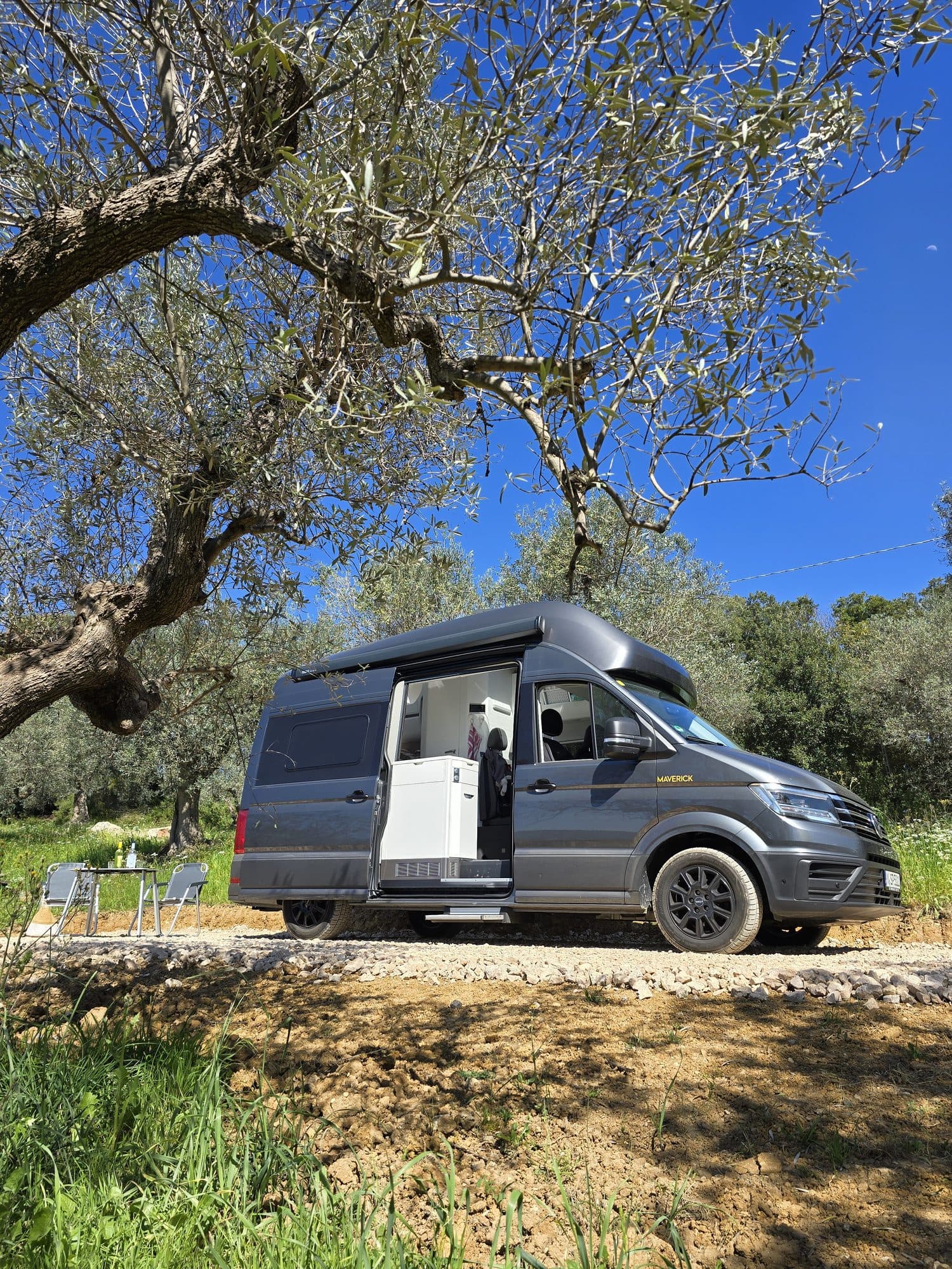 Grauer Campervan mit offener Schiebetür auf einem Kiesstellplatz unter einem alten Olivenbaum, daneben Campingtisch und Stühle
